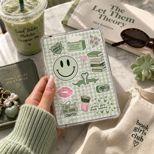 Puede incluir: Una funda transparente para tableta con un fondo de cuadros vichy verde claro decorada con varias pegatinas, incluyendo una cara sonriente grande, una pila de libros, una bebida rosa, dos dinosaurios y el texto Crying Over Fictional Characters.