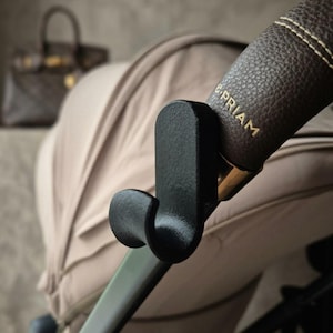 May include: A black, curved stroller hook attached to the brown, textured leather handle of a beige stroller. The handle features the gold-embossed text e-PRIAM. The hook is designed to hang items from the stroller frame securely.