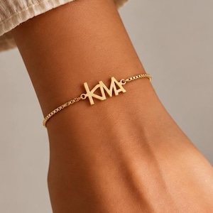 Op de afbeelding: Een goudkleurige armband met een fijne box-ketting en een centrale hanger met de letters KMA in een strak, schreefloos lettertype. De metalen bedel is aan beide zijden aan de ketting bevestigd, wat zorgt voor een gecentreerd monogramontwerp.
