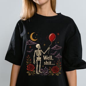 May include: A black t-shirt featuring a graphic print of a skeleton holding a red balloon. The design includes a yellow crescent moon, purple clouds, bats, and red roses. The text "Well, shit..." is printed in a light yellow font near the skeleton.