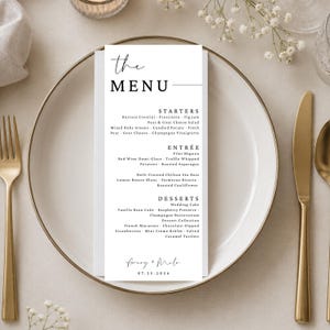 May include: A tall, rectangular white paper menu card with black text centered on a round white plate. The menu lists starters, entrees, and desserts, featuring the names Avery + Milo and the date 07.25.2026 at the bottom in a clean, modern font.