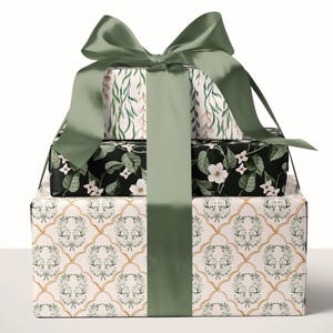 May include: A stack of three rectangular gift boxes wrapped in patterned paper, tied together with a wide sage green satin ribbon bow. The top box features a trailing leaf print, the middle box has a black floral pattern, and the bottom box displays a gold trellis design with botanical motifs.
