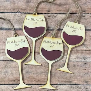 May include: Four light-colored wooden wine glass ornaments with jute twine hangers. Each ornament features a dark maroon cutout representing wine in the bowl and the engraved text "Faith n Sip" followed by the year "2023" on the upper section.
