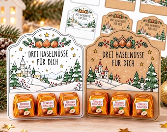 Könnte beinhalten: Zwei dekorative Kartenhalter aus Karton für drei Ferrero Küsschen Pralinen, einer in Weiß und einer in Kraftpapierbraun. Beide zeigen eine Winterlandschaft mit Schloss, Tannen und dem Text "Drei Haselnüsse für dich" sowie drei Haselnüssen.