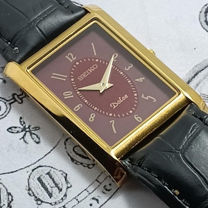 Könnte beinhalten: Eine rechteckige Seiko Dolce Armbanduhr mit goldfarbenem Gehäuse, dunkelrotem Zifferblatt mit goldfarbenen Zeigern und arabischen Ziffern von 3 bis 12. Die Uhr verfügt über ein schwarzes Lederarmband mit strukturierter Oberfläche und Krone.