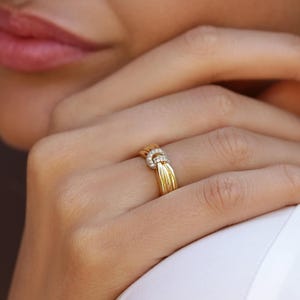 Può includere: Un anello in oro caratterizzato da un design a più fili che convergono al centro. L'elemento centrale a forma di nodo è impreziosito da piccole pietre rotonde e trasparenti incastonate a pavé sulla superficie del metallo dorato.
