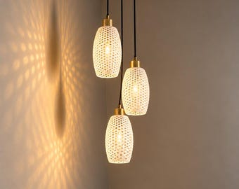 Puede incluir: Una lámpara colgante de tres luces con pantallas cilíndricas blancas con un patrón de puntos perforados. Cada pantalla cuelga de un cable negro desde un dosel de techo de latón circular, emitiendo una luz cálida que proyecta patrones.