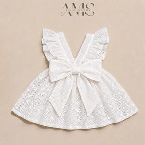 Puede incluir: Un vestido de tela de encaje blanco con tirantes con volantes y un gran lazo central en la parte delantera. La prenda tiene una falda acampanada y un escote en V. El texto AMS SEWING PATTERNS es visible en la parte superior central.