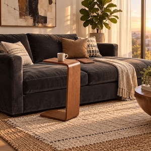 May include: A C-shaped wooden side table with a smooth finish, positioned next to a dark gray sofa. The table holds a white ceramic mug. The sofa features a brown throw pillow, a patterned accent pillow, and a cream-colored knit throw blanket.