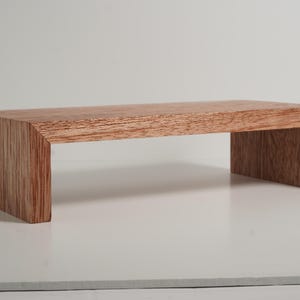 May include: A rectangular wooden riser with a natural reddish-brown wood grain finish. The stand features a flat top surface supported by two vertical side panels, creating a bridge-like structure with a clean, mitered corner design.