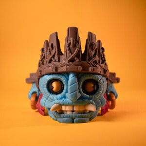 May include: A stylized collectible figure featuring a blue, mask-like face with large circular eyes, prominent white fangs, and a textured brown crown-like headpiece. The figure has small red accents on the sides and a matte finish throughout.