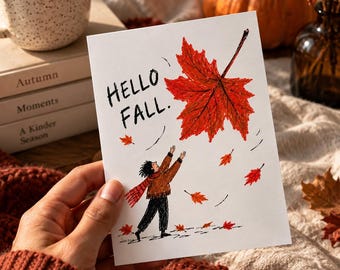 May include: A white rectangular greeting card featuring a hand-drawn illustration of a person in a brown sweater and red scarf reaching toward a large red maple leaf. The text HELLO FALL is written in black ink. Several smaller red leaves are scattered.