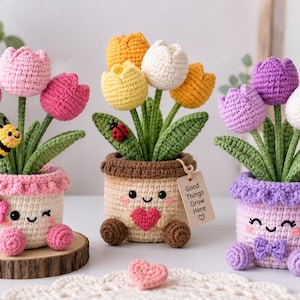 Könnte beinhalten: Drei gehäkelte Tulpenarrangements in Töpfen mit lächelnden Gesichtern. Der linke Topf ist rosa-weiß mit einer Biene, der mittlere Topf ist braun-beige mit einem Marienkäfer und einem Schild mit der Aufschrift Good Things Grow Here, der rechte Topf ist lila.