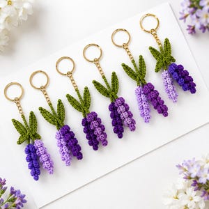 May include: Six crochet lavender flower keychains arranged in a row. Each keychain features a gold-tone metal ring and chain attached to a green crocheted stem with two leaves and two clusters of purple and light purple crocheted flower buds.