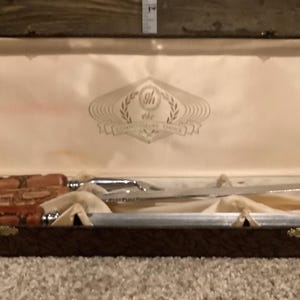 Puede incluir: Un juego de trinchar compuesto por un cuchillo largo y un afilador, ambos con mangos marrones texturizados. El conjunto se presenta en un estuche rectangular marrón con forro interior de satén color crema y un logotipo impreso en la tapa.