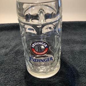 May include: A clear glass beer mug featuring a dimpled exterior texture and a sturdy handle. The front displays the Erdinger Weissbräu logo in blue, red, and white, with the text Erdinger Weissbier printed clearly below the circular emblem.