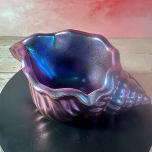 May include: A seashell-shaped bowl featuring a metallic, iridescent finish that shifts between deep purple, blue, and magenta tones. The surface has a subtle glittery texture, and the bowl displays a spiraled, ridged exterior with a wide, open mouth.