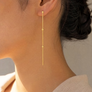 Può includere: Un lungo e sottile orecchino a catena color oro, caratterizzato da un filo metallico delicato impreziosito da tre piccole sfere dorate distanziate uniformemente. L'orecchino pende verticalmente dal lobo, creando una silhouette lineare ed essenziale.