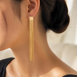 Puede incluir: Un pendiente largo de tono dorado que presenta una pequeña pieza superior cuadrada con un acabado de metal cepillado y texturizado. A la base cuadrada se une una cascada de múltiples cadenas finas y delicadas que cuelgan verticalmente.