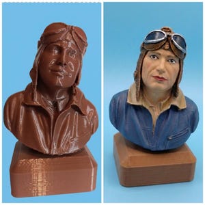 Può includere: Un confronto fianco a fianco di un busto di pilota stampato in 3D. Il busto a sinistra è in plastica marrone monocromatica. Il busto a destra è dipinto a mano con una giacca blu, colletto beige, cuffia da aviatore in pelle marrone e occhiali.