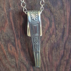 May include: A metallic pendant necklace featuring a tapered, rectangular charm with embossed floral and geometric patterns. The charm is suspended from a simple, thin metal chain link, showcasing a polished finish with intricate detailing.