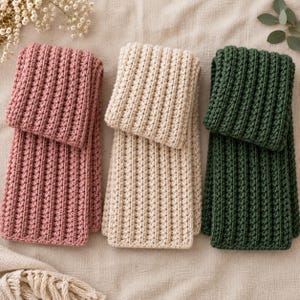 Pode incluir: Três cachecóis retangulares com um padrão de malha canelada vertical. Os cachecóis são apresentados em três cores sólidas distintas: um rosa empoeirado, um branco creme e um verde floresta profundo. Cada cachecol está dobrado no topo.