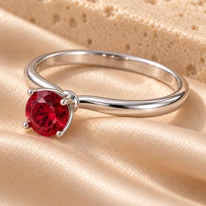 May include: A silver-toned metal ring featuring a single round-cut vibrant red gemstone held in a four-prong setting. The band is smooth and polished, showcasing a simple design that highlights the central red stone as the primary focal point.