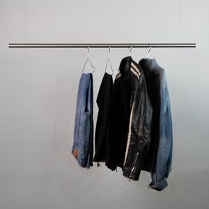 Könnte beinhalten: Eine horizontale, von oben hängende Kleiderstange aus Metall mit vier Drahtkleiderbügeln. Daran hängen eine blaue Jeans, ein schwarzes Kleidungsstück, eine schwarze Lederjacke mit weißen Streifen und eine blaue Jeansjacke.