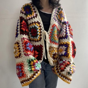 Pode incluir: Um cardigã de manga comprida feito de quadrados de crochê multicoloridos em tons de vermelho, amarelo, azul, verde e roxo, unidos por fio cor de creme. A peça apresenta um fecho frontal de amarrar com dois cordões de fio cor de creme.