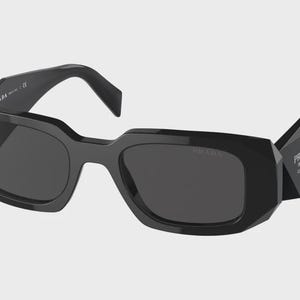 Può includere: Occhiali da sole neri con una montatura spessa, angolare e geometrica e lenti scure. Le aste presentano una forma triangolare prominente con il logo Prada Milano stampato in bianco. Il marchio Prada è visibile anche sull'asta interna.