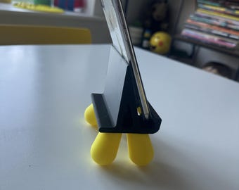 May include: A black smartphone stand with four rounded yellow feet holding a silver smartphone in an upright position. The stand features a triangular support structure and a grooved base to secure the device, providing a stable platform for viewing.