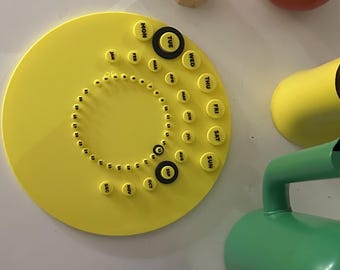May include: A circular yellow perpetual calendar featuring black text for days of the week, months, and numbers one through thirty-one. The calendar includes three black circular markers for tracking dates, surrounded by a small toy figure and pitchers.