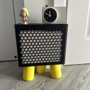 Peut inclure: Une enceinte rectangulaire noire avec une grille frontale en nid d'abeille et trois pieds cylindriques jaune vif. Une petite figurine blonde et une horloge analogique blanche avec un cadran noir et des aiguilles blanches sont posées dessus.