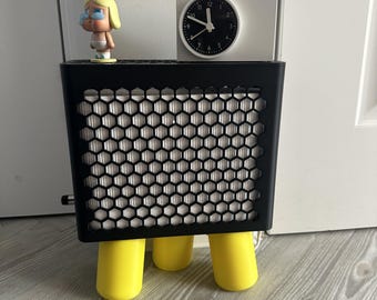 May include: A black rectangular air purifier with a honeycomb front grille and three bright yellow cylindrical legs. On top sits a small white analogue clock with a black face and a small blonde figurine wearing a white nappy.
