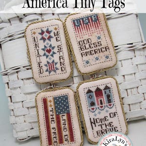 Könnte beinhalten: Vier rechteckige, kreuzgestickte Anhänger mit patriotischen Themen. Jeder Anhänger hat einen goldfarbenen Rand und eine kleine Metallschlaufe zum Aufhängen. Die Anhänger zeigen amerikanische Flaggen, Sterne und Sätze wie "God Bless America".