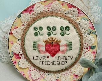 Peut inclure: Une broderie au point de croix circulaire présentant un motif Claddagh avec un cœur rouge, une couronne dorée et des trèfles verts sur un tissu crème. Le texte indique LOVE, LOYALTY, FRIENDSHIP. Monté sur une assiette florale avec dentelle.