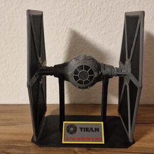Pode incluir: Um modelo de caça estelar TIE/LN cinza e preto montado num suporte de exposição preto. O cockpit esférico apresenta uma janela circular com uma moldura hexagonal. Uma placa retangular na base exibe o texto TIE/LN em preto sobre um fundo amarelo.