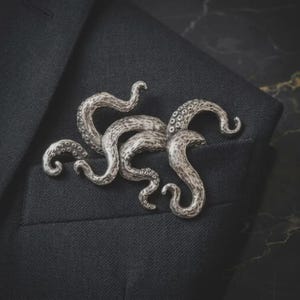 May include: A silver-toned metal brooch shaped like a cluster of octopus tentacles. The piece features a textured surface with visible suction cup details along the curved, winding arms. The brooch is pinned to a dark charcoal grey fabric garment.