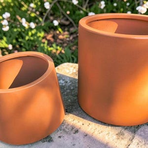 May include: Two cylindrical terracotta planters with a smooth, matte finish. The set includes one smaller, slightly tapered pot and one larger, straight-sided pot, both featuring a warm, earthy orange color and a simple, open-top design.