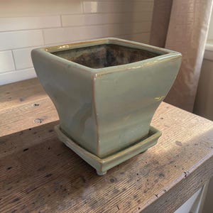 May include: A square ceramic planter with a glossy light sage green glaze and a matching square saucer base. The planter features a flared rim, slightly curved sides, and a dark, unglazed interior. The saucer has small feet at each corner.