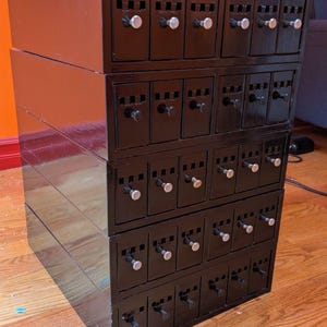 May include: A black wooden storage cabinet featuring five stacked horizontal sections, each containing six vertical drawers. Every drawer is equipped with a small silver knob and a rectangular cutout at the top for labeling or gripping purposes.