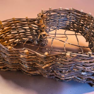 May include: A heart-shaped basket constructed from dark brown woven twigs and branches. The structure features a rustic, open-weave design with vertical stakes protruding along the top rim and a grid-like base made of thin, flexible wooden branches.