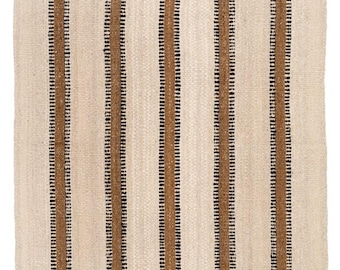 May include: A rectangular woven area rug featuring a repeating pattern of vertical stripes in shades of cream, tan, and black. The rug has a textured, natural fiber appearance with a small brown rectangular tag located in the bottom right corner.