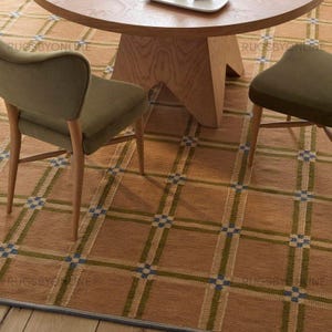 Può includere: Un tavolo rotondo in legno con finitura marrone chiaro poggia su un tappeto color cuoio con un motivo a griglia di quadrati verde oliva e blu. Due sedie imbottite color verde oliva con gambe in legno sono posizionate attorno al tavolo.