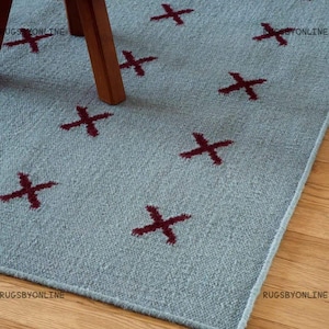 Puede incluir: Una alfombra de color azul claro con un patrón repetitivo de símbolos de cruz de color rojo oscuro. La alfombra tiene una superficie tejida texturizada y está parcialmente cubierta por las patas de madera de un mueble. El texto RUGSBYONLINE es visible.