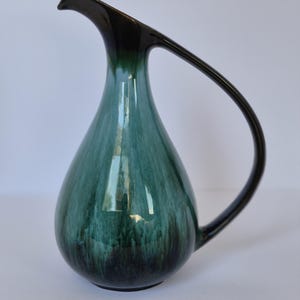 May include: A ceramic pitcher with a teardrop-shaped body and a long, curved handle. The glaze features a gradient transition from a deep, dark brown at the rim and base to a vibrant, mottled teal and forest green in the centre of the vessel.