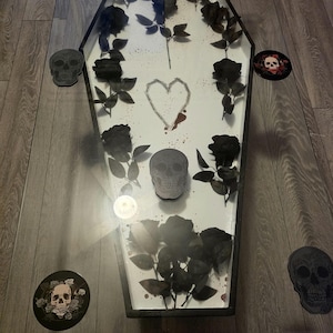 May include: A coffin-shaped coffee table with a glass top and a white interior base. The interior features black artificial roses, a central skull, a hand-drawn heart outline, and red paint splatters. Several skull-themed coasters surround the table.