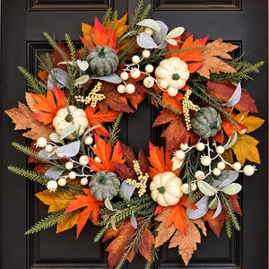 May include: A decorative autumn wreath featuring a mix of orange, red, and brown maple leaves, interspersed with small green and cream-colored pumpkins, sprigs of green pine needles, and clusters of small white berries on thin, branching stems.