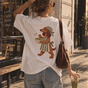 Może przedstawiać: Biały t-shirt z krótkim rękawem z grafiką brązowego jamnika w zielono-biały fartuch w paski i chustę na głowie, mieszającego miskę zielonej matchy. Koszulka jest zestawiona z niebieskimi dżinsami i brązową zamszową torbą na ramię.