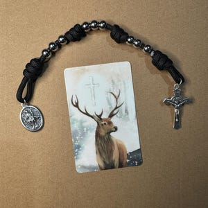 May include: A black paracord rosary featuring silver-toned metal beads, a silver crucifix, and a silver oval medal depicting a saint. The rosary is positioned above a rectangular prayer card featuring an illustration of a deer with antlers.